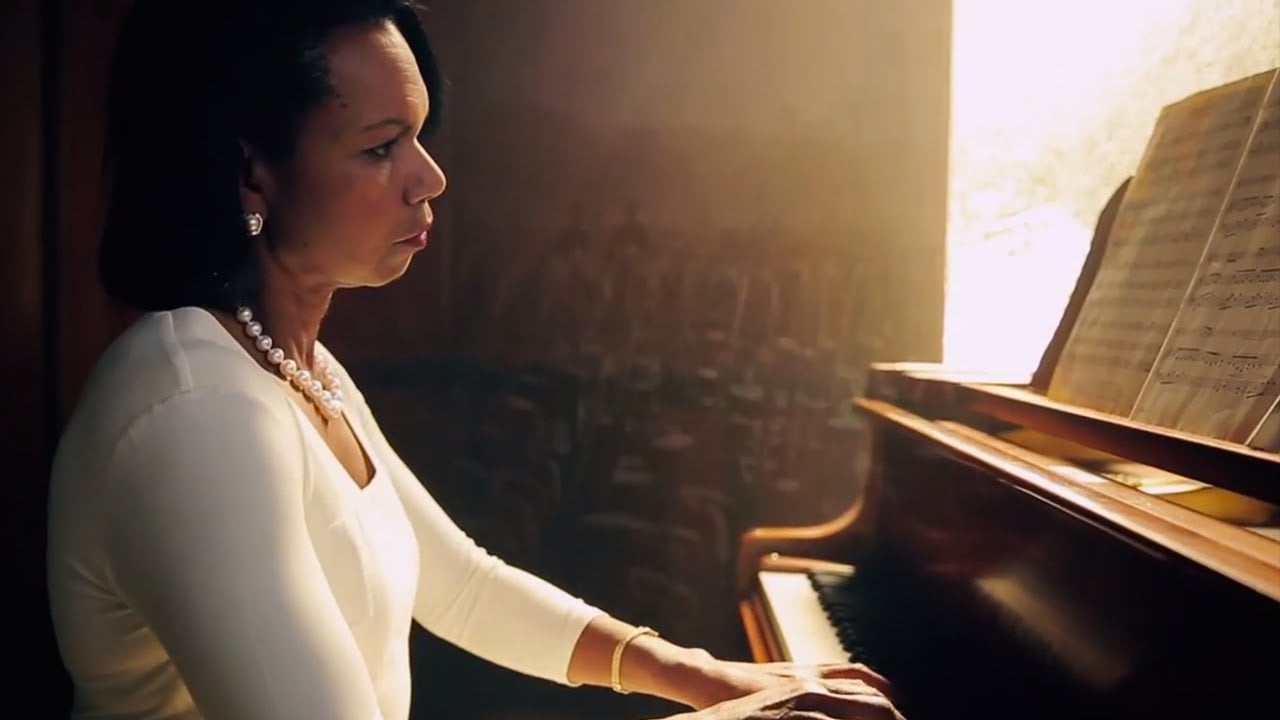 A woman in a white top and pearl necklace plays the piano, reading sheet music in a softly lit room.