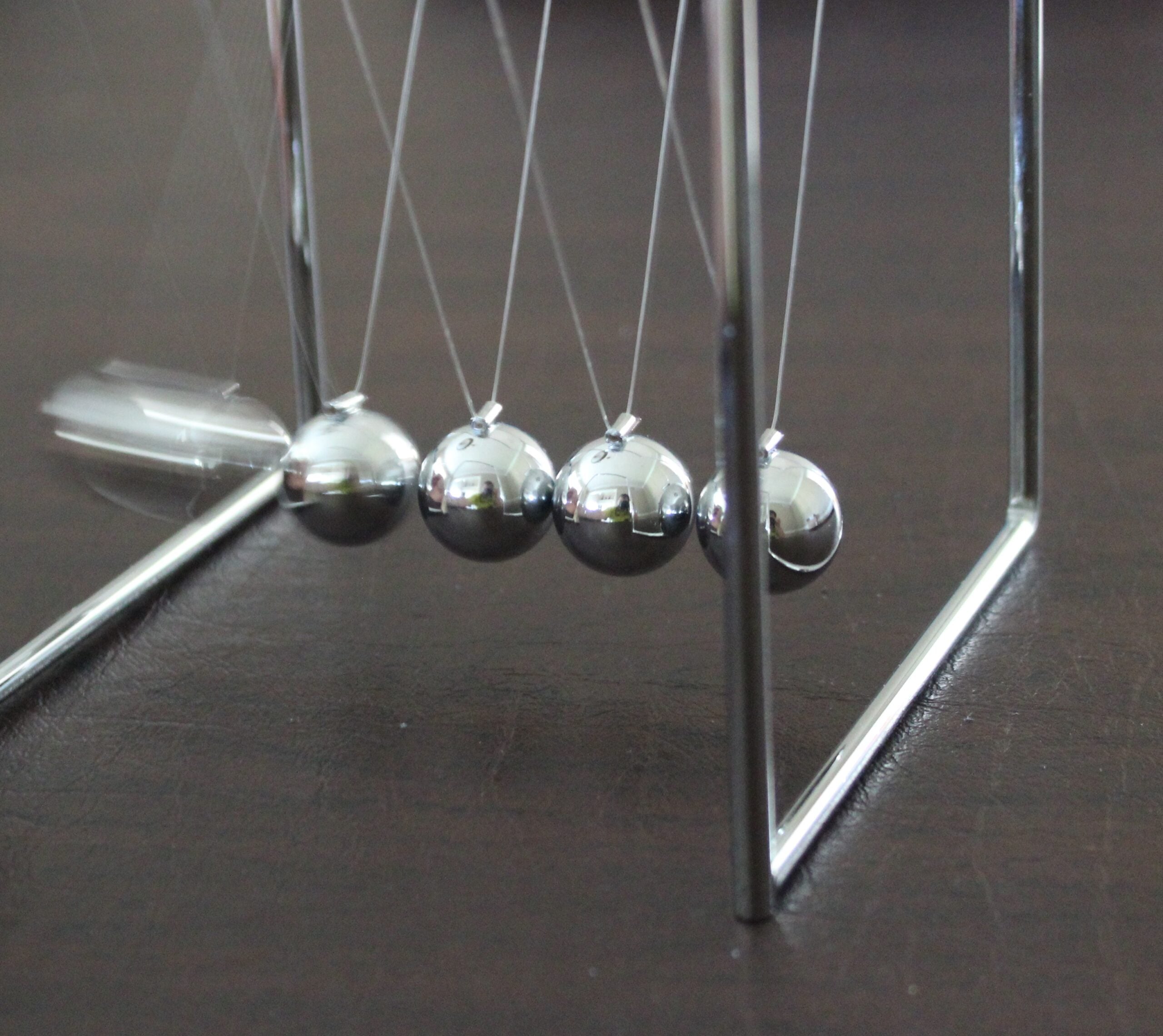 A Newton's cradle with five metal balls suspended on strings. One ball is lifted and released, showing motion blur, while the others remain still, illustrating the transfer of kinetic energy.