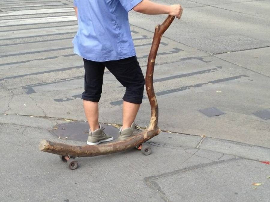 A person rides a homemade scooter with a large tree branch as the frame and handle, standing on a paved street near a crosswalk.