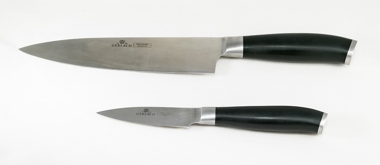 Two kitchen knives with black handles lying on a white surface, one large chef's knife and one smaller paring knife, both with shiny metal blades.