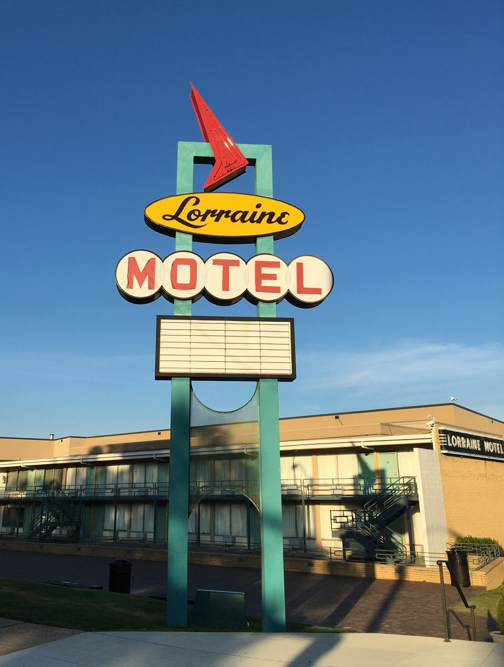 Vintage Lorraine Motel sign with red arrow, yellow and white background, and empty marquee. The motel building is visible in the background under a clear blue sky.