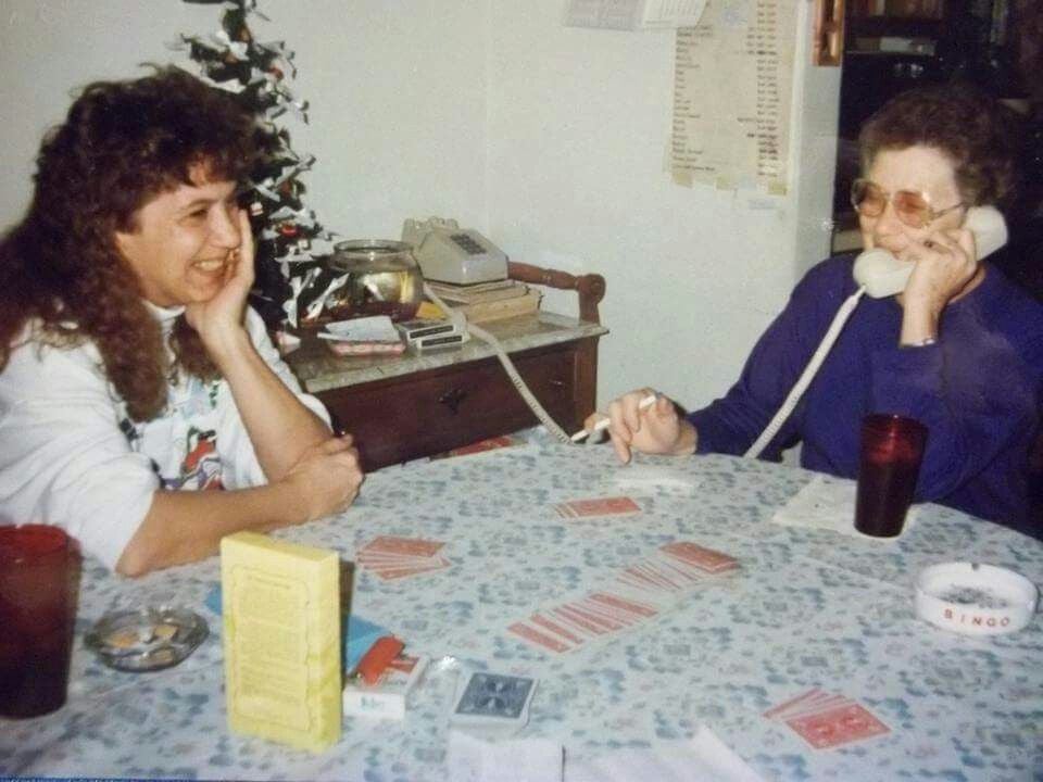 Two women sit at a table playing cards. One woman smiles while the other talks on a corded phone. There are drinks, cards, and a bingo set on the table. A small Christmas tree and telephone are in the background.