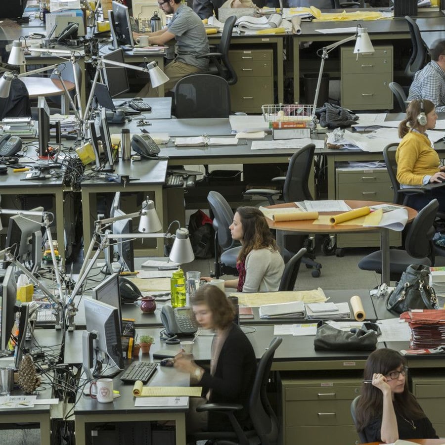 Open-plan office with several people working at desks with computers, papers, and desk lamps. The workspace is busy, with employees focused on their tasks. Some are seated, typing or talking on phones.