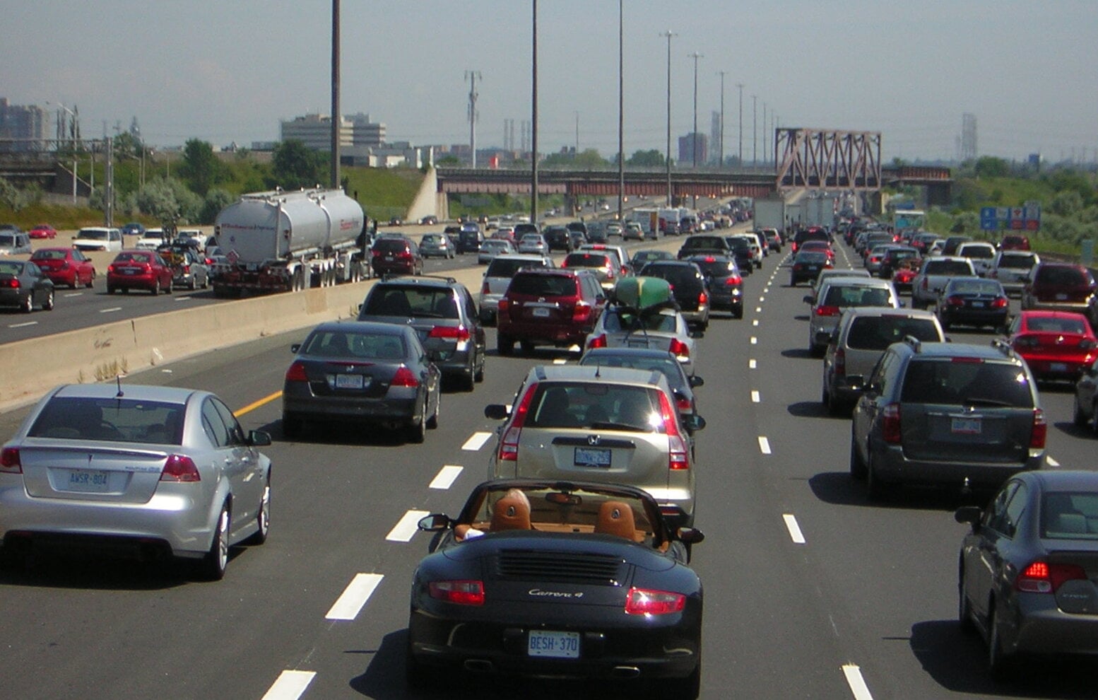 Heavy traffic fills multiple lanes on a wide highway during daytime, with cars, SUVs, and trucks in both directions. A bridge and city skyline are visible in the background under a clear sky.