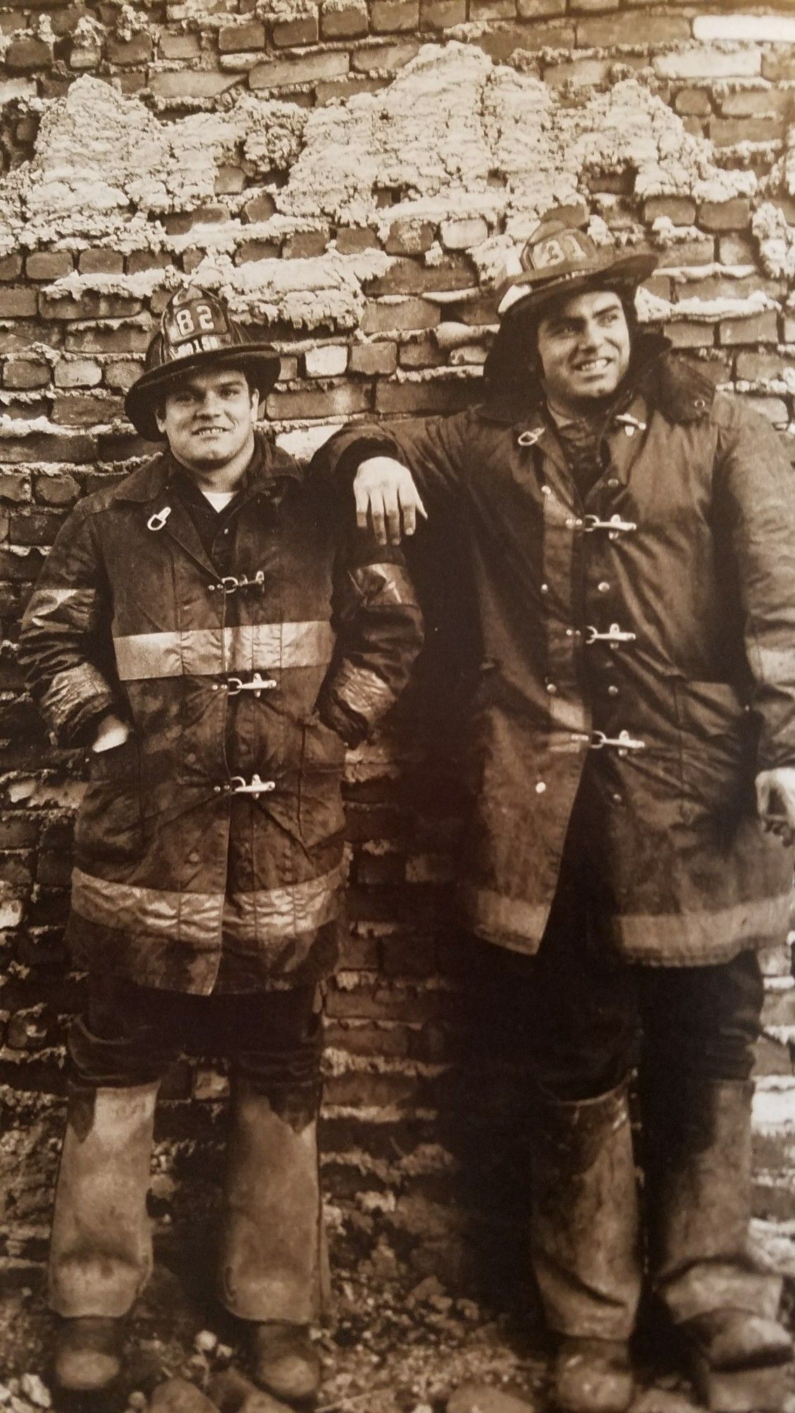Two firefighters in old-fashioned gear stand side by side against a weathered brick wall. One has his arm resting on the other's shoulder. Both are smiling, and their helmets display the number 82.