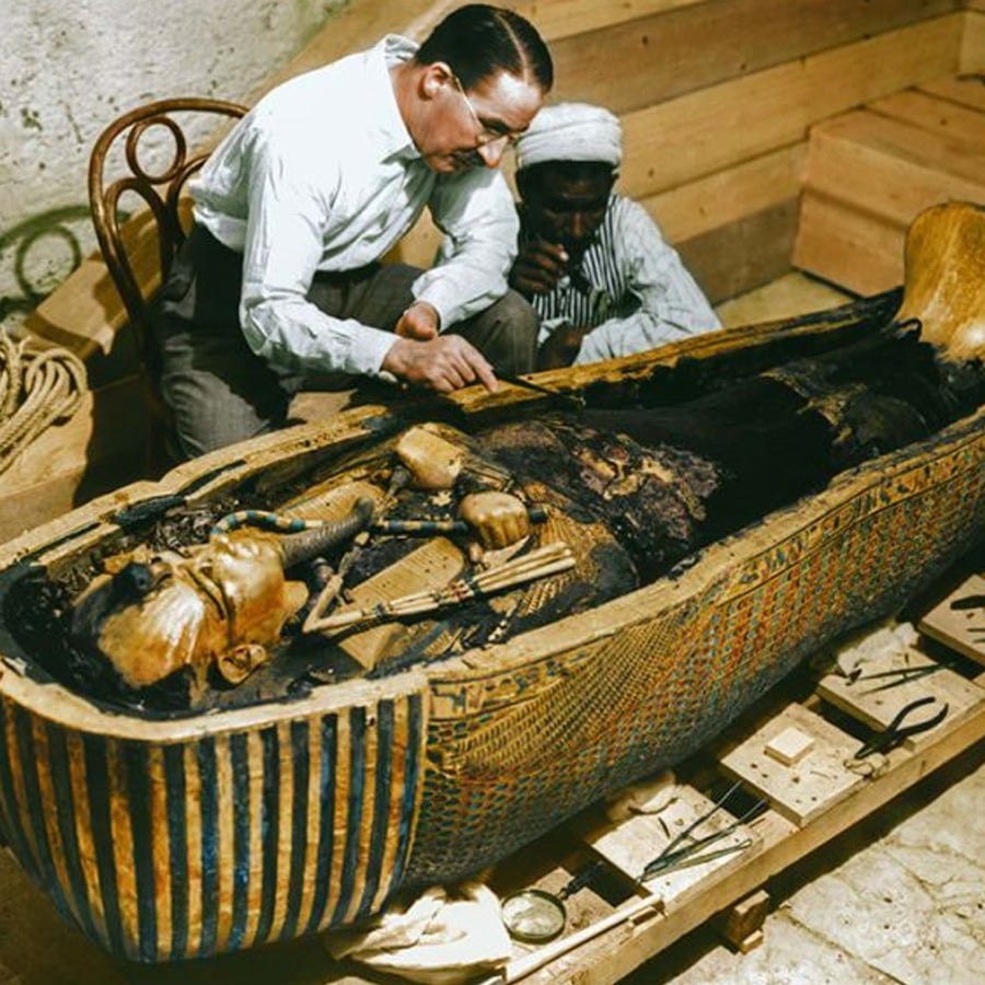 Two men examine the ornate, partially opened sarcophagus of an Egyptian mummy in a wooden room, surrounded by tools and artifacts.