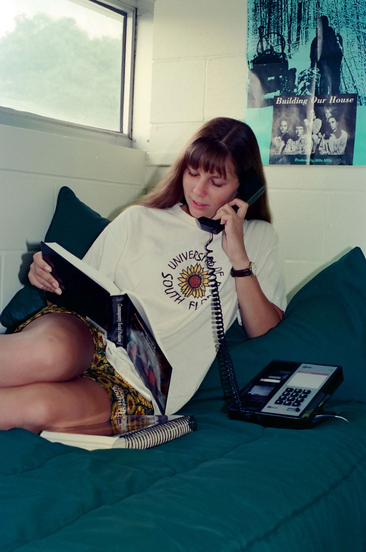 A young woman sits on a bed, talking on a corded phone while reading a book. A notebook and a phone rest beside her, and a poster is on the wall behind her.