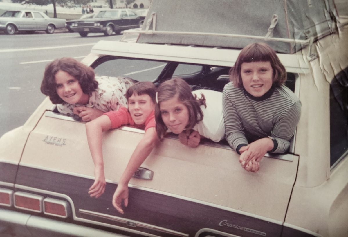 Four children are lying on the open tailgate of a vintage beige car, smiling at the camera. The car is parked by a street, and two classic cars are visible in the background.