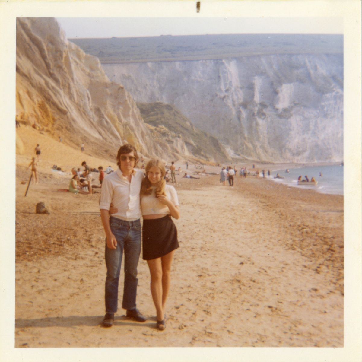 A young couple stands arm in arm on a sandy beach with cliffs in the background. Other people are scattered along the shore and swimming in the sea on a sunny day.