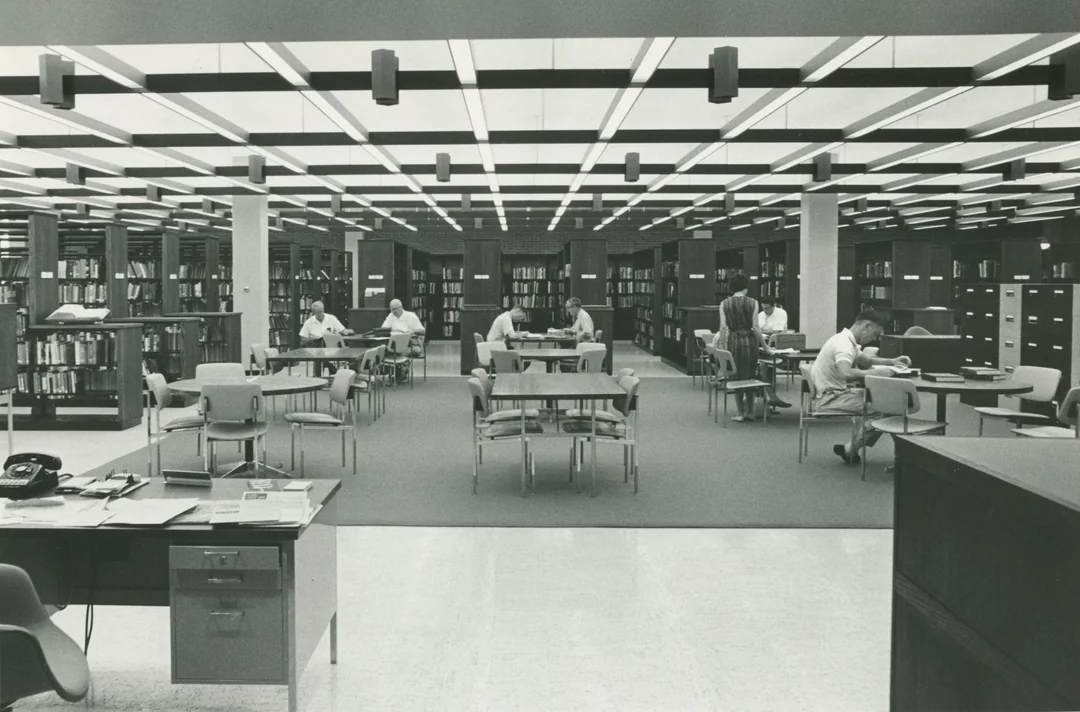 Black-and-white photo of a spacious library with people reading at tables, surrounded by bookshelves and filing cabinets, under a grid-patterned ceiling with fluorescent lights.