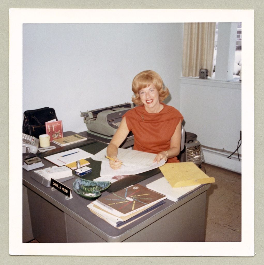 A woman with blonde hair and a red top sits smiling at a desk in an office, holding a pencil over papers. The desk has folders, a typewriter, a nameplate, and office supplies. Large windows are in the background.