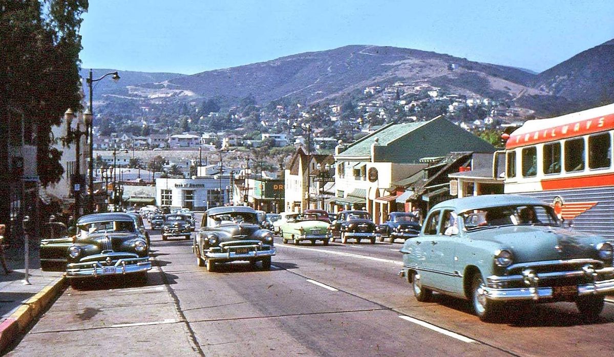 A busy street scene from the 1950s shows classic cars driving past shops and buildings, with hills and houses in the background under a clear blue sky.