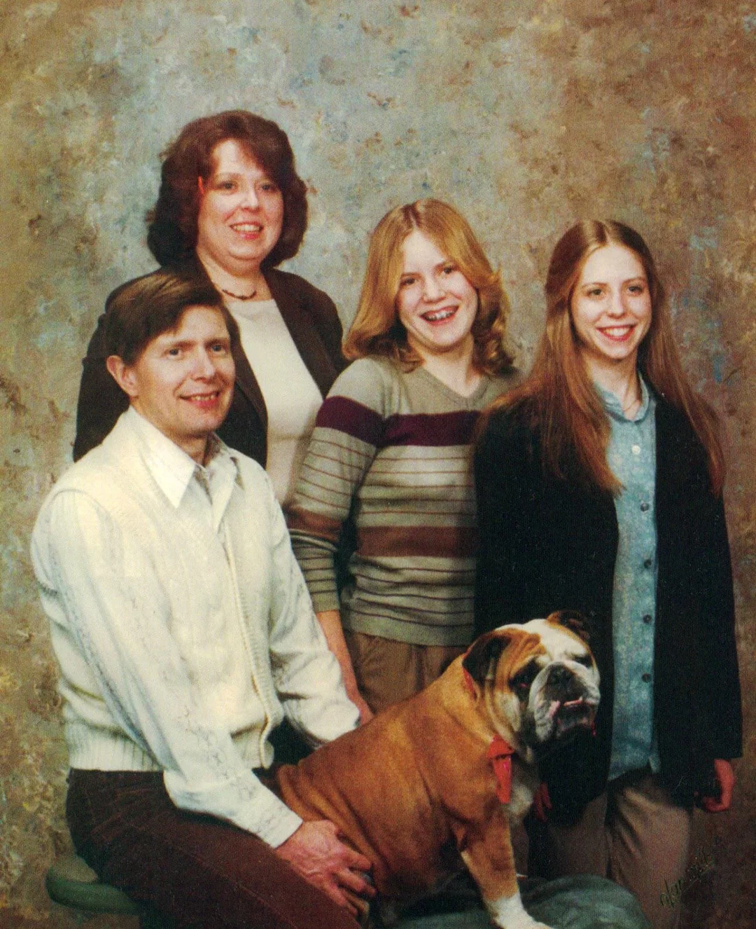 A vintage family portrait of three women, one man, and a bulldog. Everyone is smiling and wearing 1970s-style clothes, posing in front of a mottled studio backdrop.