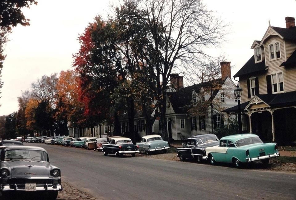 A street lined with vintage cars parked along old houses with gabled roofs, surrounded by large trees with autumn leaves. The scene appears to be from the mid-20th century.