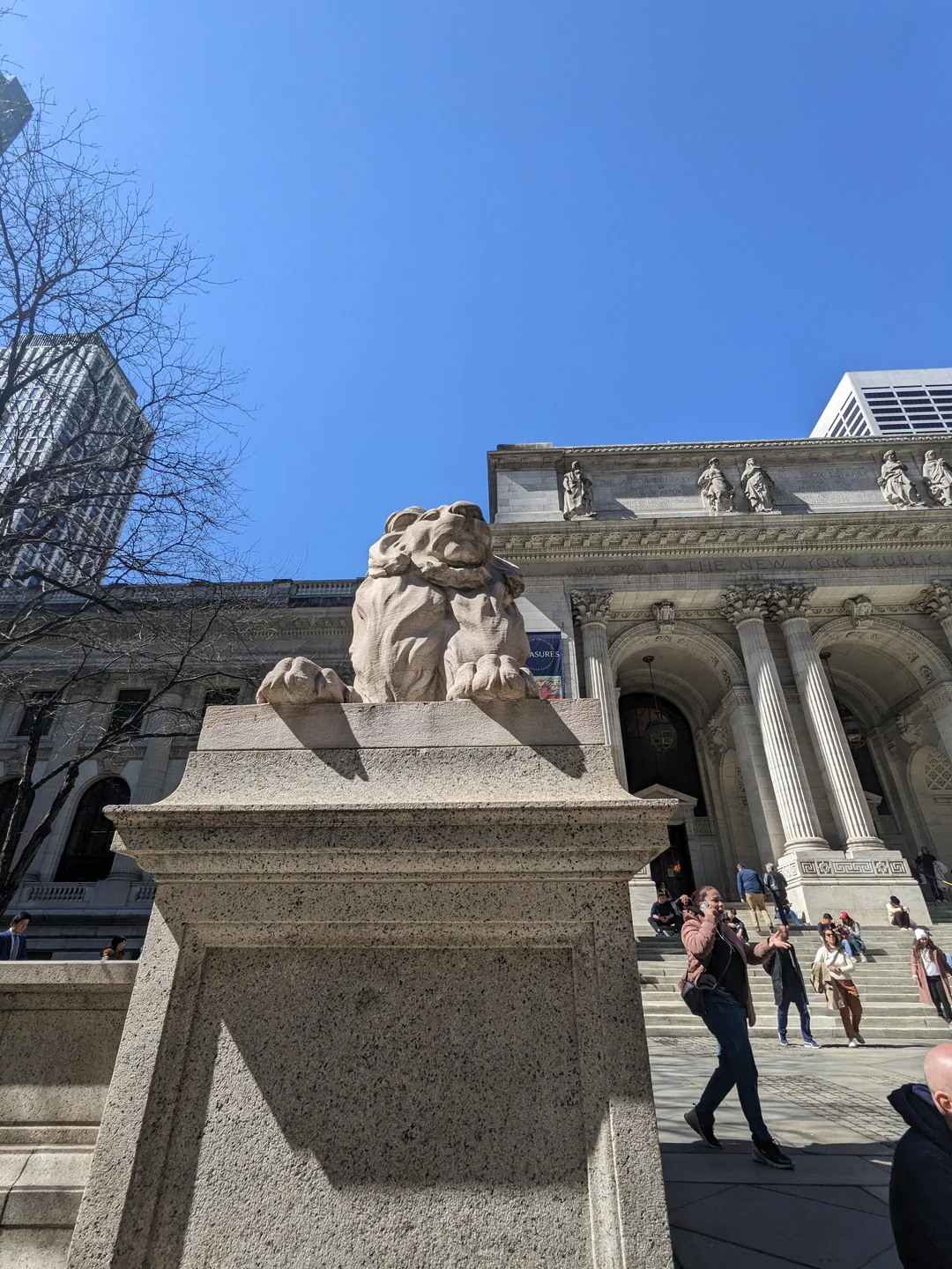 A stone lion statue sits atop a pedestal outside a large, classical building with columns and arches, as people walk and sit on the steps under a clear blue sky.