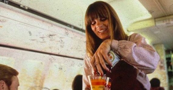 A smiling flight attendant serves drinks to passengers on an airplane, pouring from a can into a plastic cup. The cabin has patterned red seats, and sunlight streams through the windows.