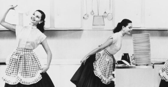 Three women in matching vintage dresses and aprons pose cheerfully in a kitchen, each standing near a tall stack of dishes on a counter, with utensils and cabinets in the background.