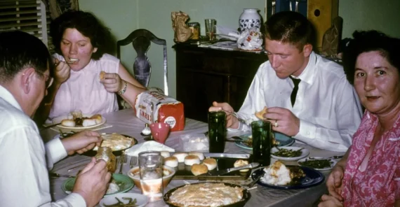 Four adults sit around a dining table enjoying a meal with pie, biscuits, green beans, and drinks. The table is covered with a gray cloth, and the room has a cozy, retro atmosphere.