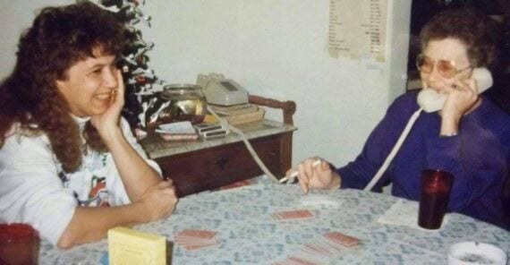 Two women sit at a table playing cards. One smiles while the other talks on a corded phone. A small Christmas tree and old-style phone are in the background. A Bingo ashtray, red cups, and card game accessories are on the table.
