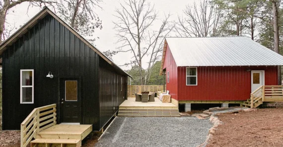 Two small houses, one black and one red, sit side by side with wooden steps, porches, and a connecting deck in a wooded area with bare trees and gravel pathway.