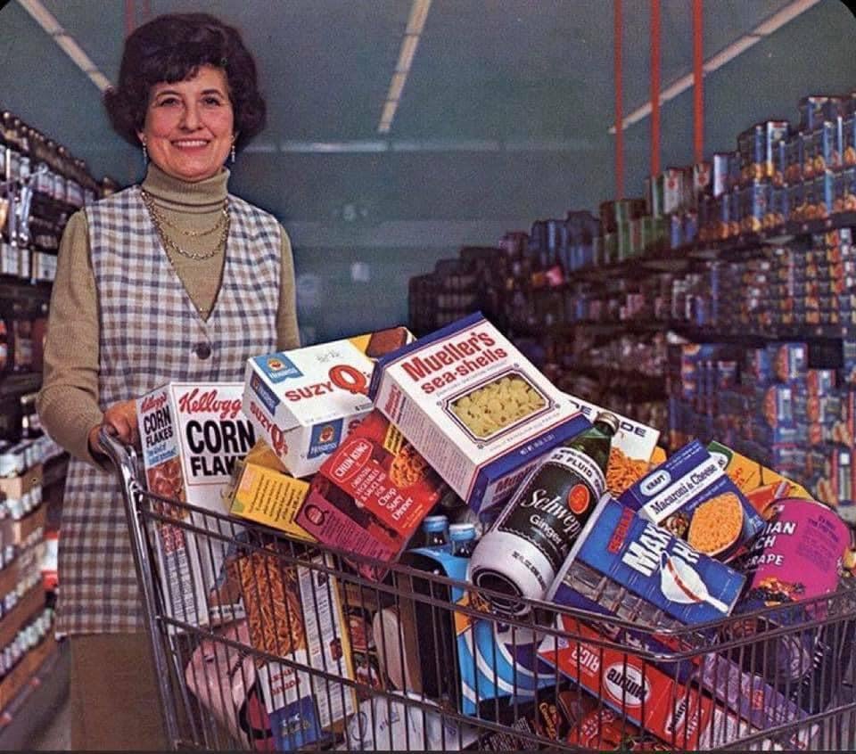 A woman smiles while standing in a grocery store aisle, pushing a shopping cart overflowing with various boxed and canned food items, including cereal, pasta, crackers, and canned goods.
