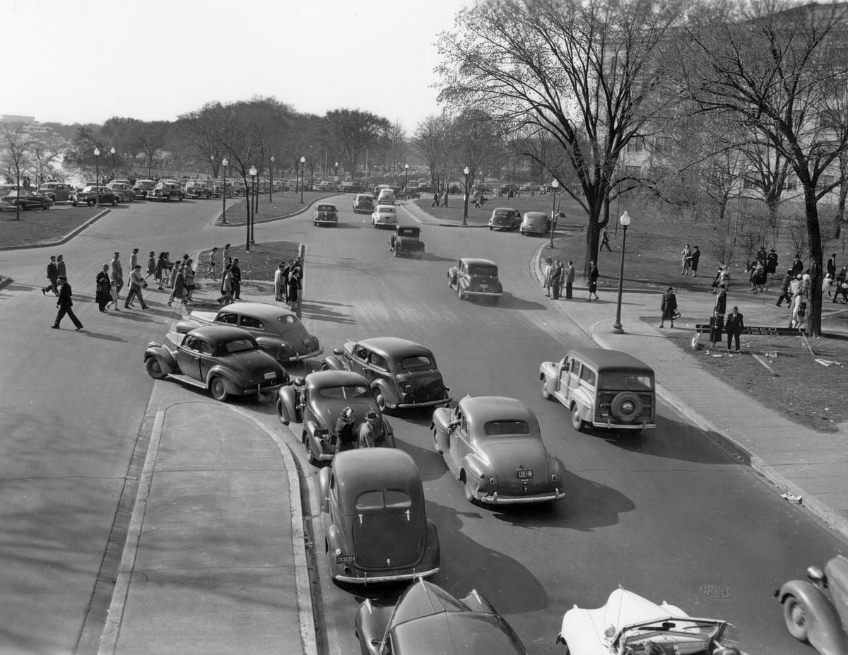 Black-and-white photo of a busy street scene from the 1940s, with vintage cars parked and driving, pedestrians crossing, and people walking along tree-lined sidewalks.