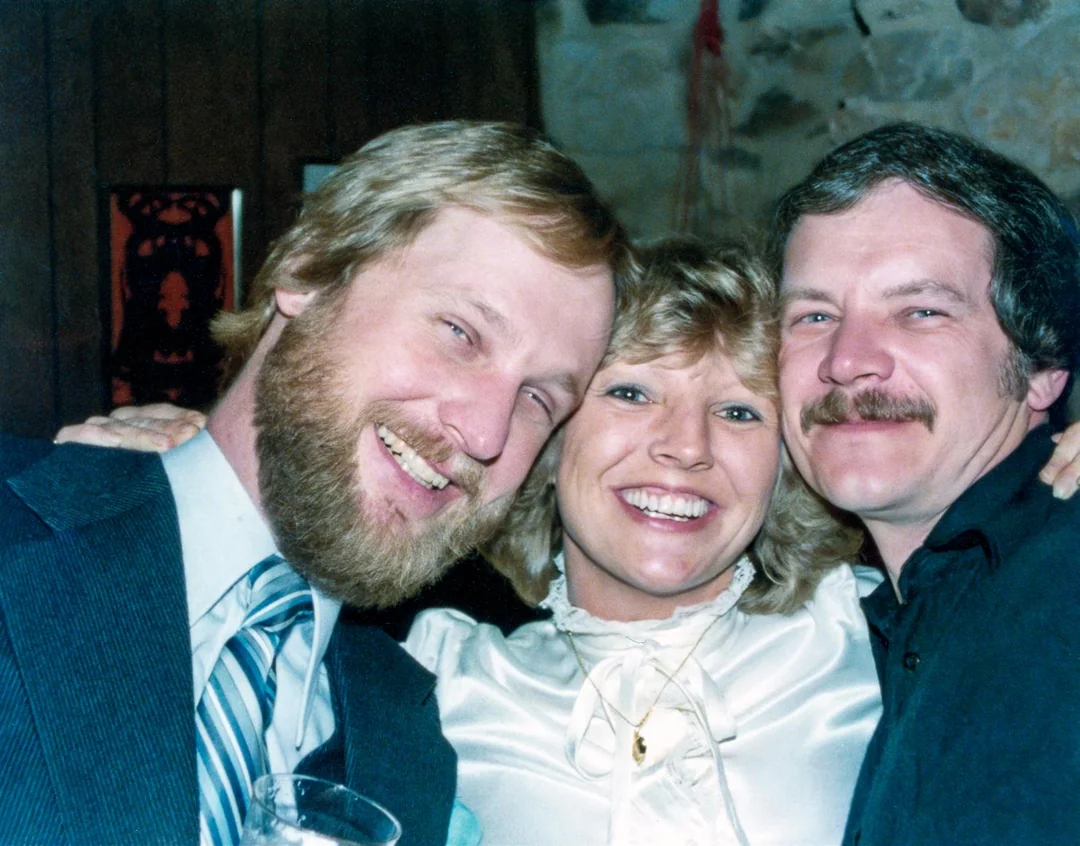Three adults smiling and hugging closely together at an indoor gathering. The man on the left has a beard, the woman in the center has blonde hair, and the man on the right has a mustache.