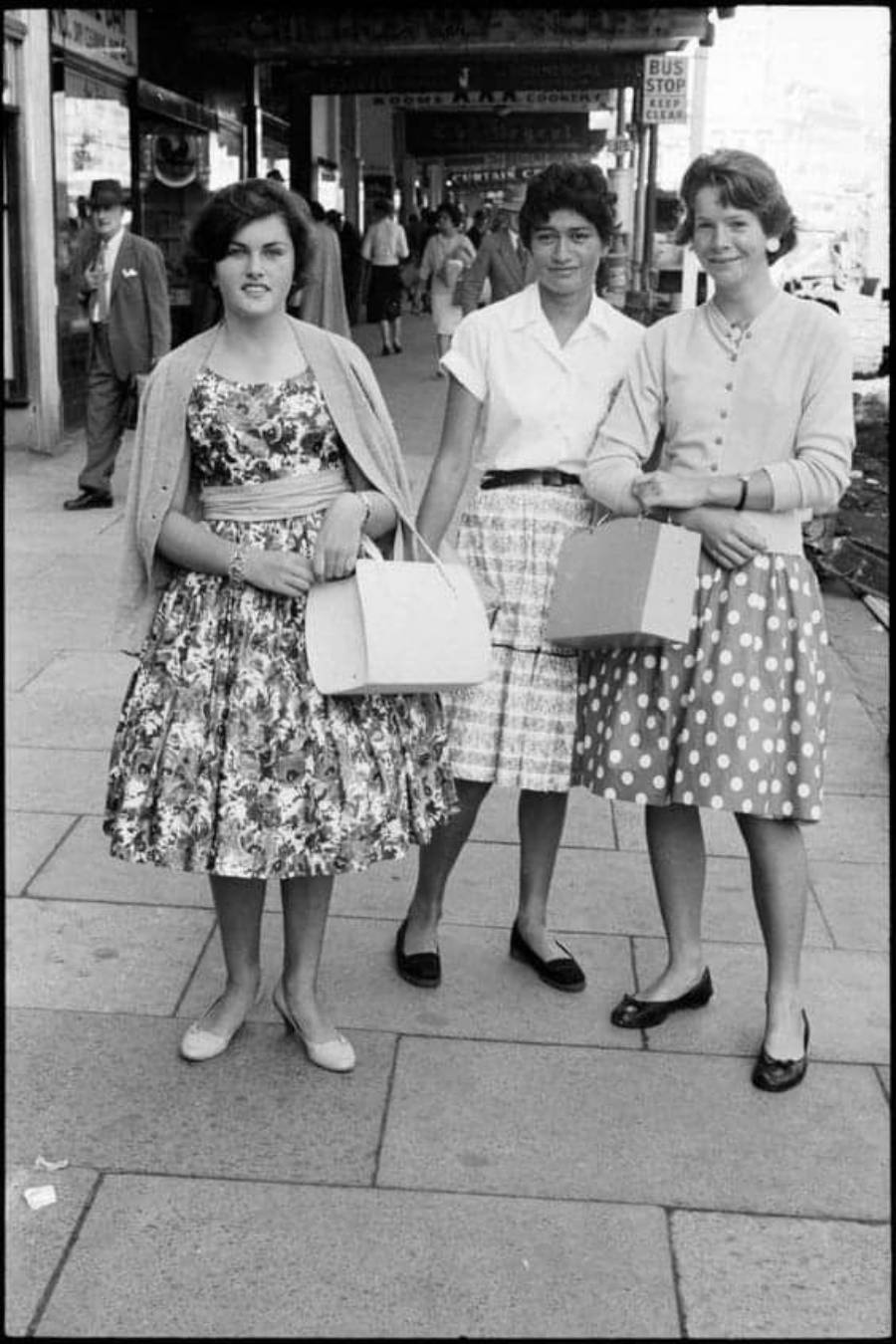 Three young women in 1950s-style dresses and cardigans stand on a city sidewalk, each holding a handbag. People and shops line the street in the background.