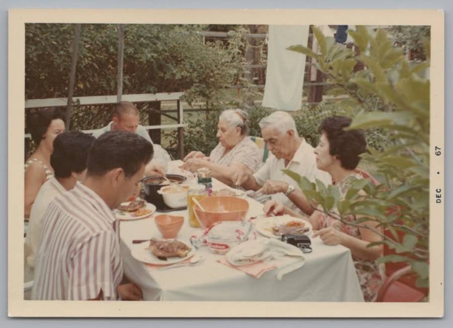 Six adults sit around a table outdoors, sharing a meal. The table holds various dishes, and the group appears to be enjoying a casual gathering. Trees and greenery surround the scene. Photo dated December 1967.