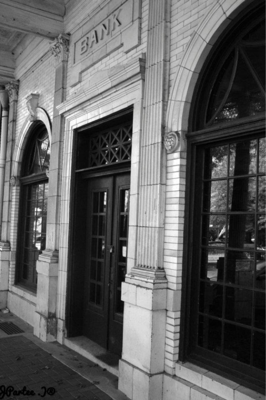 Black-and-white photo of an old brick building with arched windows and a doorway. The word "BANK" is engraved above the door. Trees and reflections are visible through the windows.