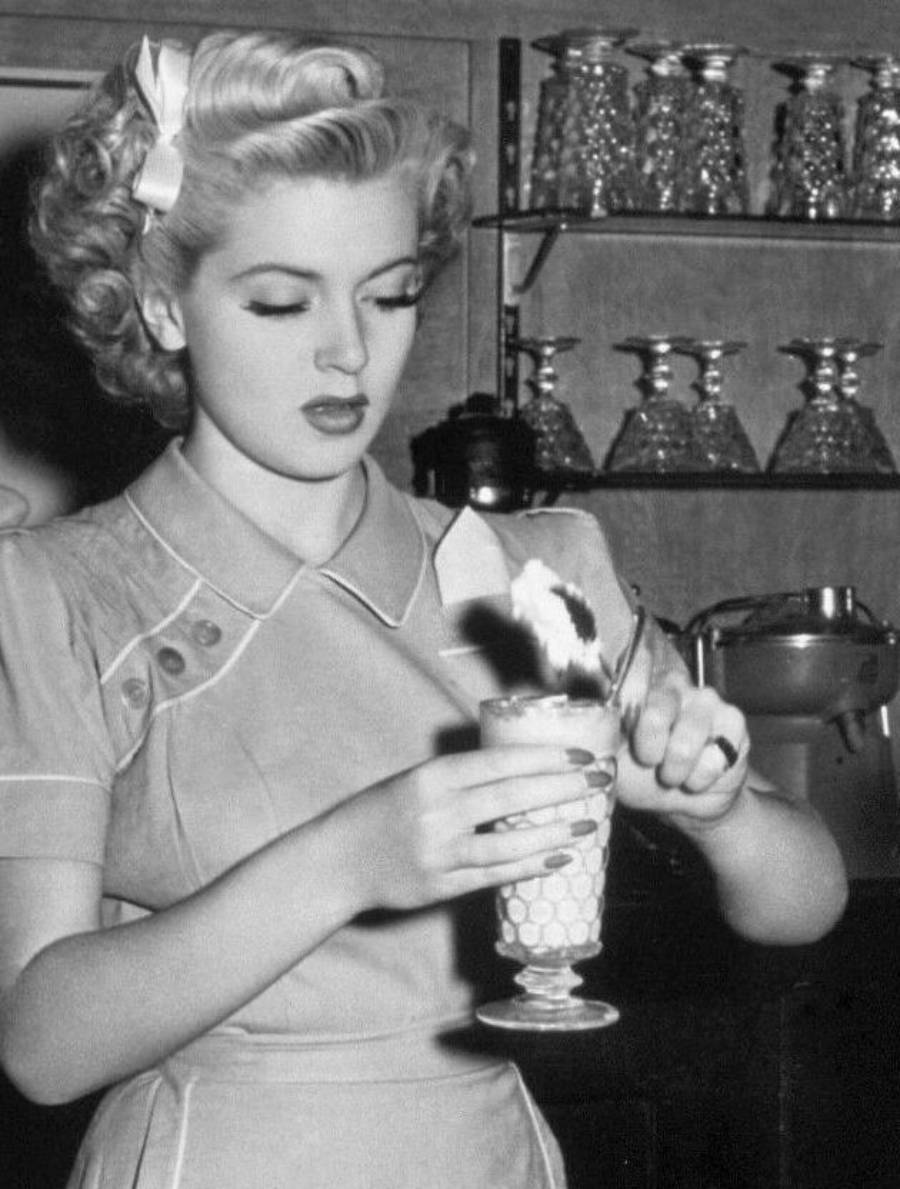 A woman in vintage attire holds a tall glass and scoops ice cream into it. She stands in front of shelves with glasses, with her hair curled and wearing a dress with buttons and a bow in her hair.