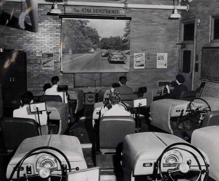 Students sit in car simulator desks facing a large screen showing a road scene, participating in a driving lesson in a classroom labeled “Aetna Drivotrainer.”