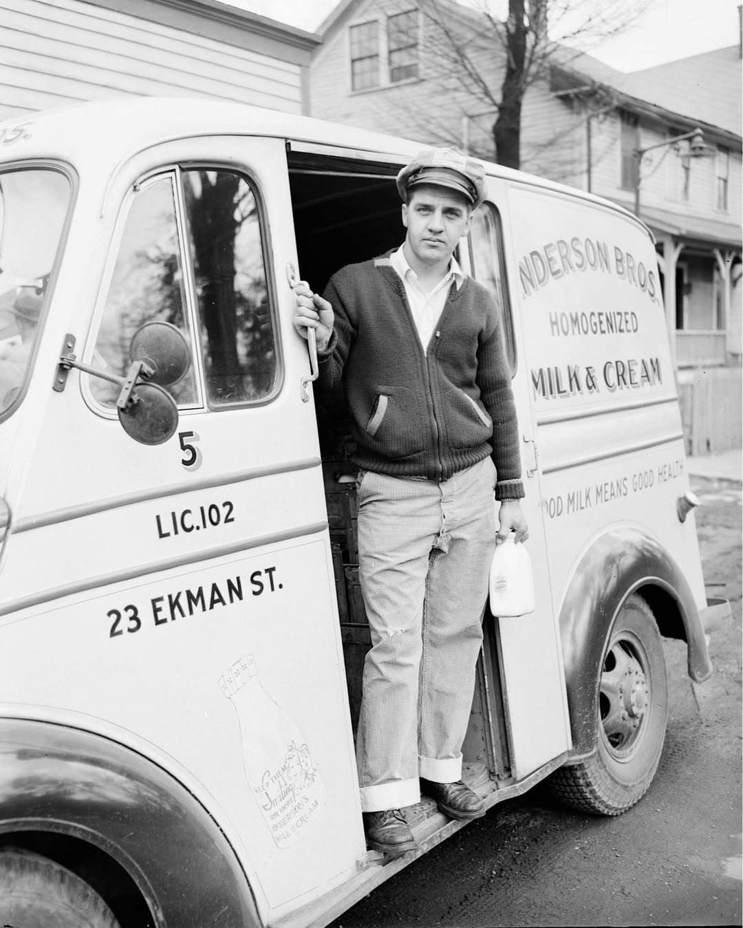 A milkman in a cap and cardigan stands at the open door of an Anderson Bros. milk truck, holding a bottle. The truck is parked on a residential street beside houses.