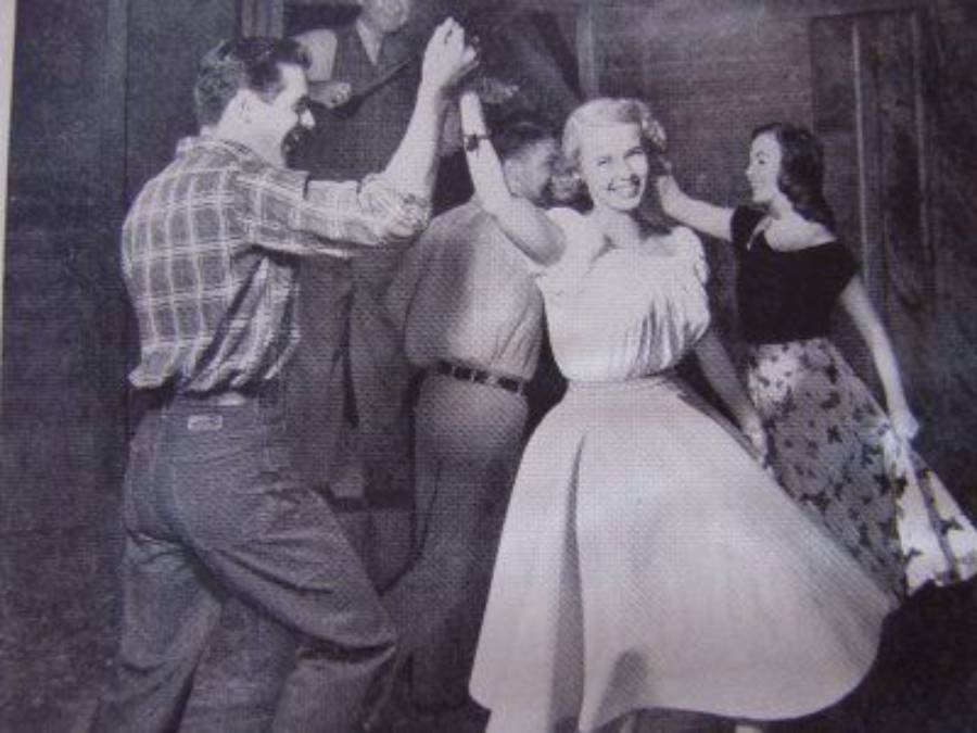 Four people are dancing together in a lively scene. The women wear skirts, and one spins with a smile on her face. The men wear collared shirts and slacks. The image is in black and white.