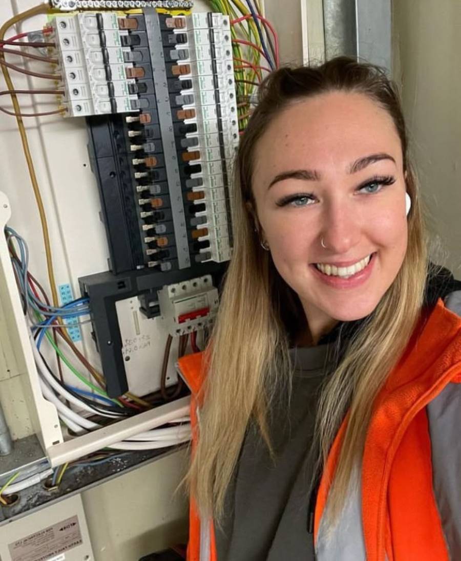 A woman wearing an orange safety jacket smiles at the camera while standing next to an open electrical panel with many wires and switches.