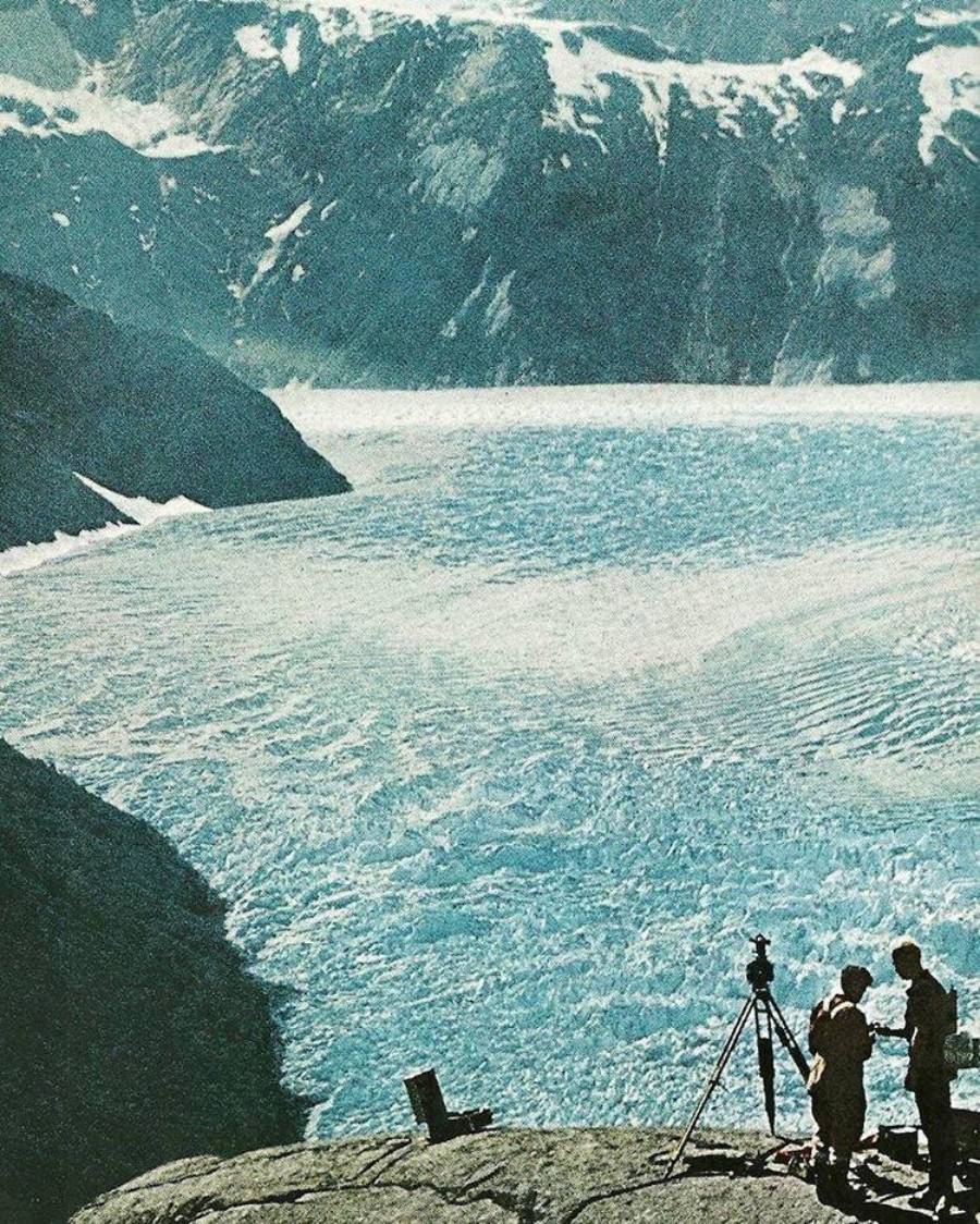 Two people stand on a rocky ledge with a camera on a tripod, overlooking a vast glacier and snow-capped mountains in the distance under a clear sky.