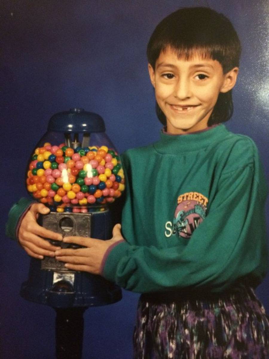 Smiling child with short dark hair holding a large gumball machine. The child wears a teal sweatshirt and patterned bottoms, standing against a blue background.