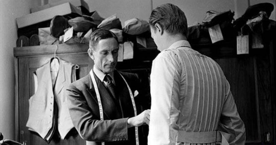 A tailor in a suit measures a client's chest with a tape measure in a fitting room. Various vests and jackets are hanging in the background.