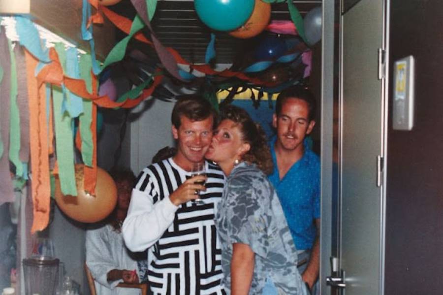 Three adults at a lively party with colorful streamers and balloons. One man holds a drink, while a woman kisses his cheek. Another man stands behind them, all smiling and enjoying the festive atmosphere.