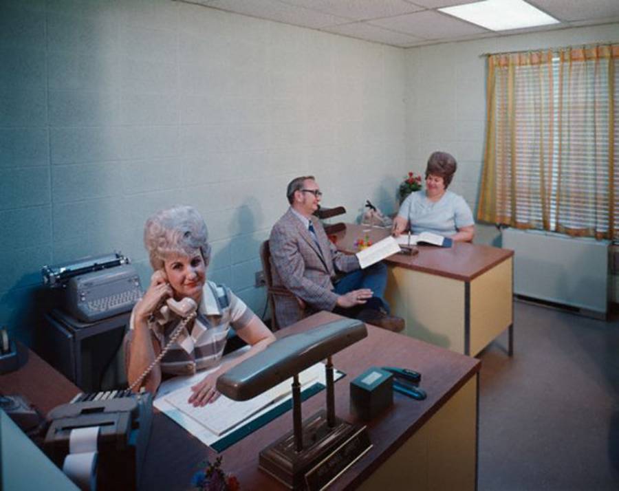 Three people in a vintage office: a woman with gray hair speaks on a corded phone at her desk, while a man in a suit and another woman talk at a nearby desk. The room has pale walls, old-fashioned decor, and a typewriter.