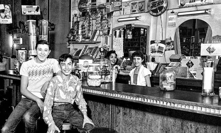 Black and white photo of two boys sitting at a diner counter, smiling at the camera. Two girls stand behind the counter, also smiling. The diner is decorated with signs, mirrors, and various items on shelves.