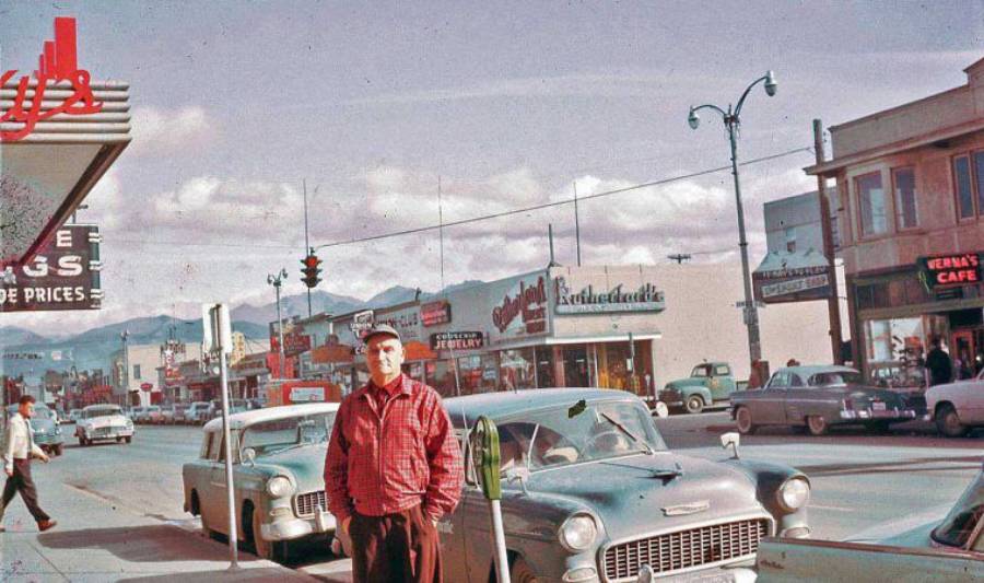 A man in a red plaid shirt stands on a busy 1950s city street lined with vintage cars and storefronts, including a cafe and liquor store, with mountains visible in the background.