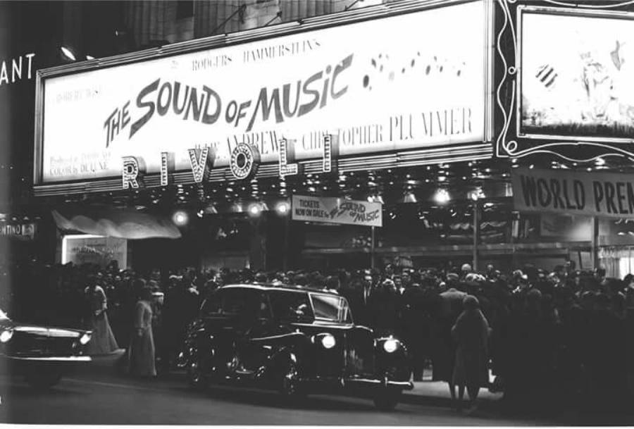 A crowd gathers outside the Rivoli Theatre at night for the world premiere of "The Sound of Music." Classic cars are parked in front, and the brightly lit marquee displays the film's title and stars.