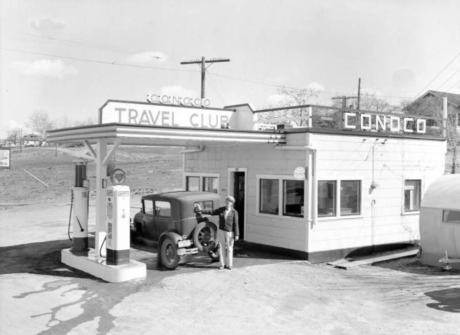 A vintage black-and-white photo of a Conoco gas station with a car at the pump and a person standing nearby. The building has signs reading "Travel Club" and "Conoco." The scene appears from the early 20th century.