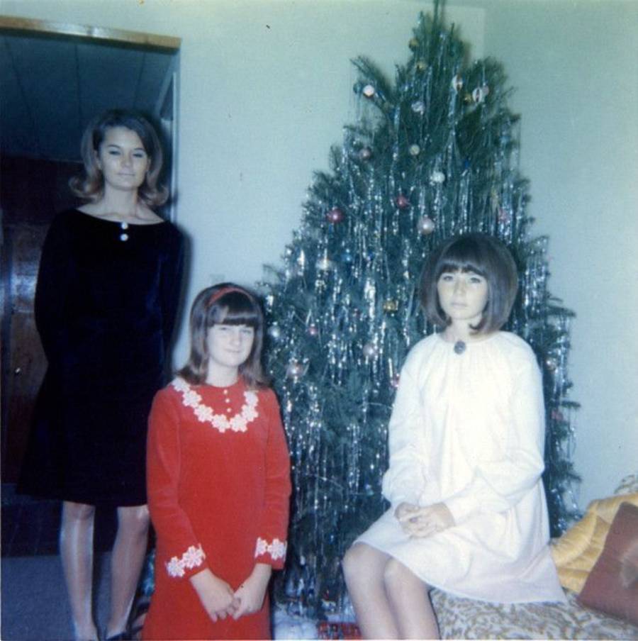 Three girls in vintage dresses pose in front of a decorated Christmas tree in a living room. One girl is sitting, while the other two are standing and smiling. The tree is adorned with tinsel and ornaments.
