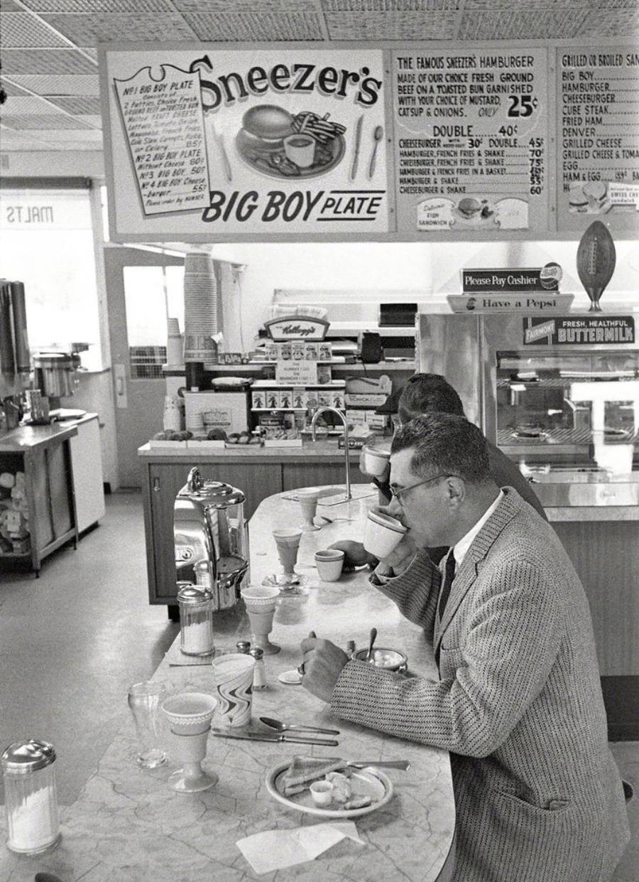 Two men in suits sit at a diner counter drinking coffee. Behind them, a menu board advertises burgers and pies. The scene has a retro, vintage feel with classic diner decor and counter seating.
