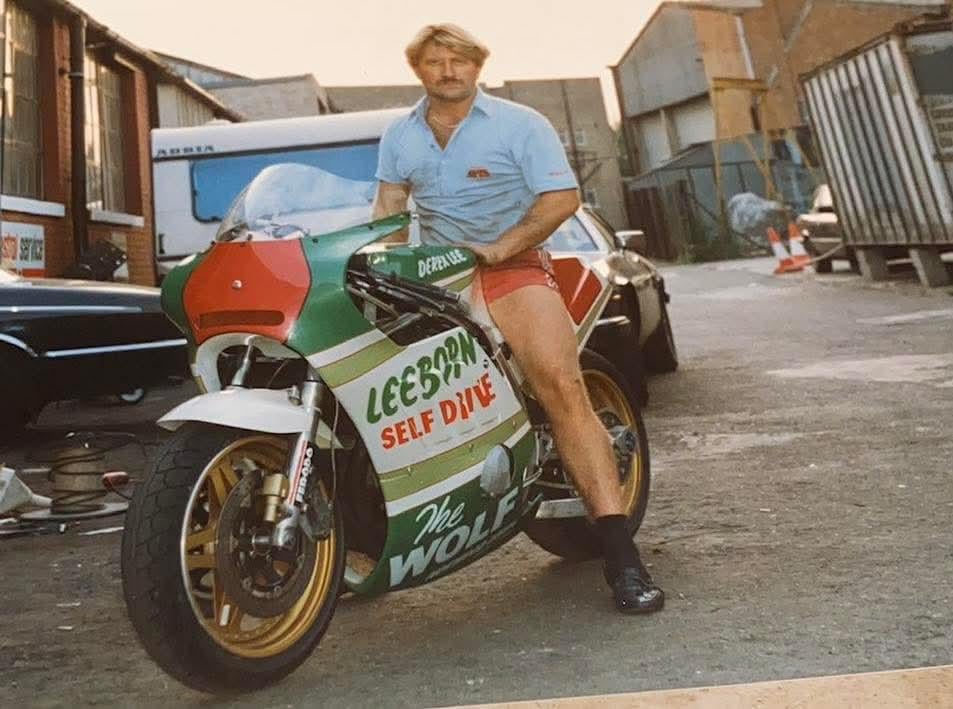 A man in shorts and a light blue shirt sits on a green, white, and red racing motorcycle labeled "The Wolf" in an alley with cars and industrial buildings in the background.