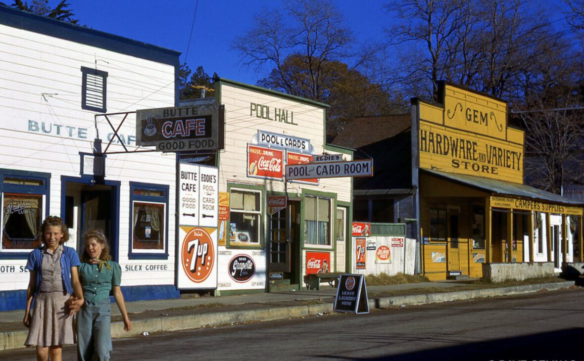 Two women walk past vintage storefronts, including a cafe, pool hall, and hardware store, on a small-town street lined with colorful signs and old-fashioned advertisements.