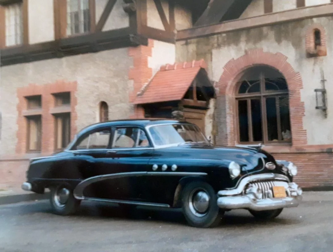 A vintage black car with chrome trim is parked in front of a building with arched windows and brick accents, evoking a classic, historical feel.