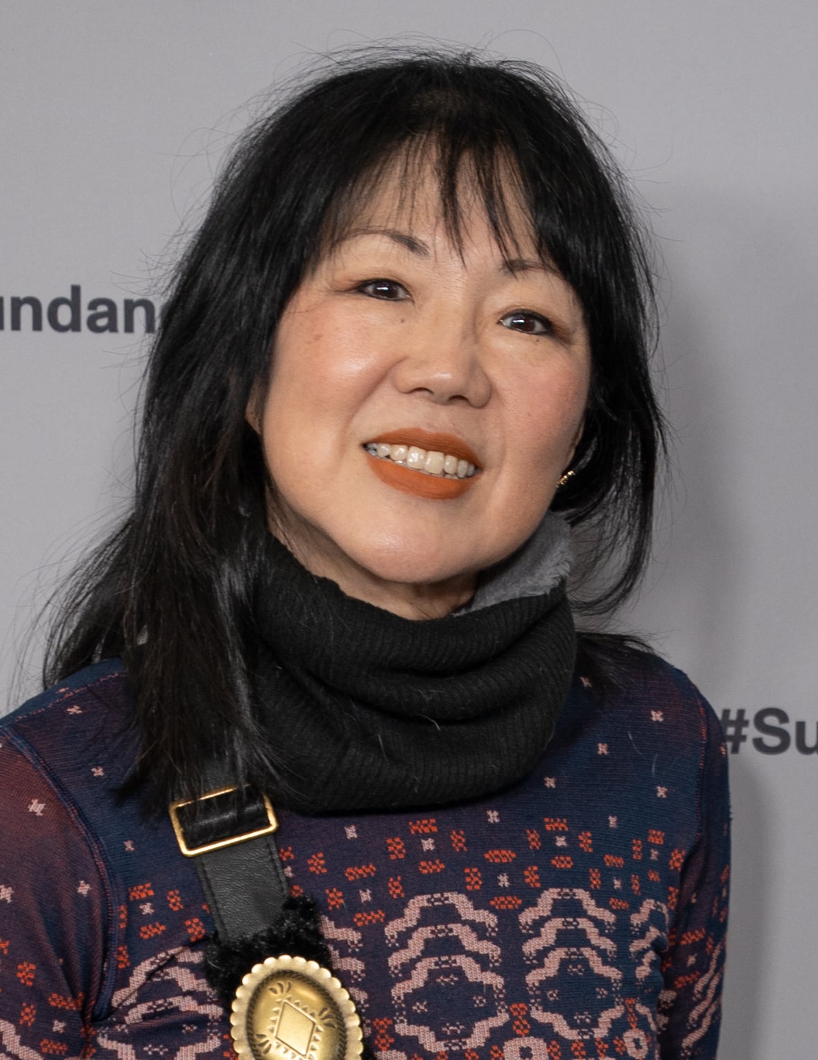 A woman with straight black hair and bangs smiles at the camera. She is wearing a patterned top, a black scarf, and a round gold pin, standing in front of a gray background with partial text visible.